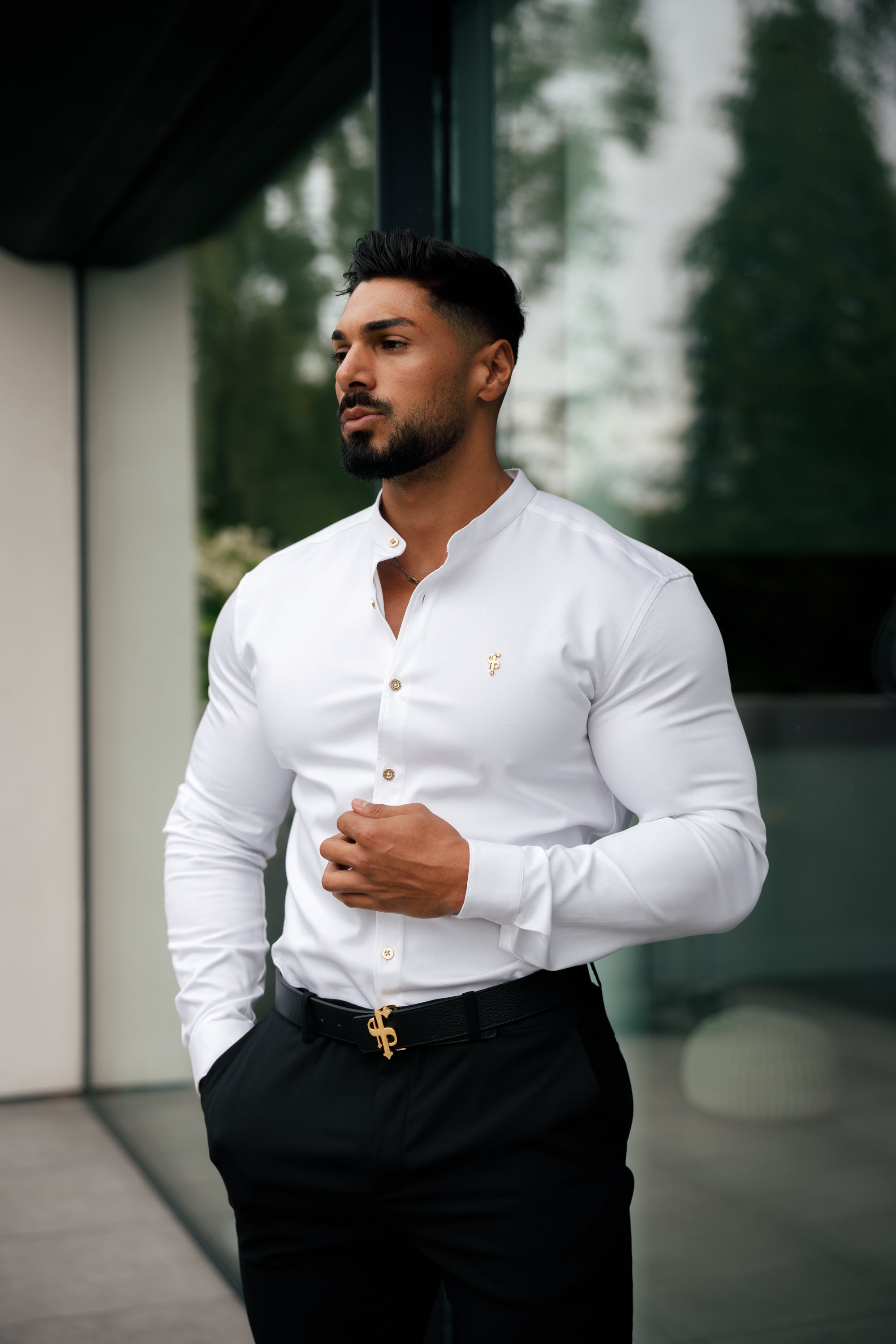 A man wearing a white long sleeve denim shirt with metal buttons and a Grandad collar, standing in front of a glass building.