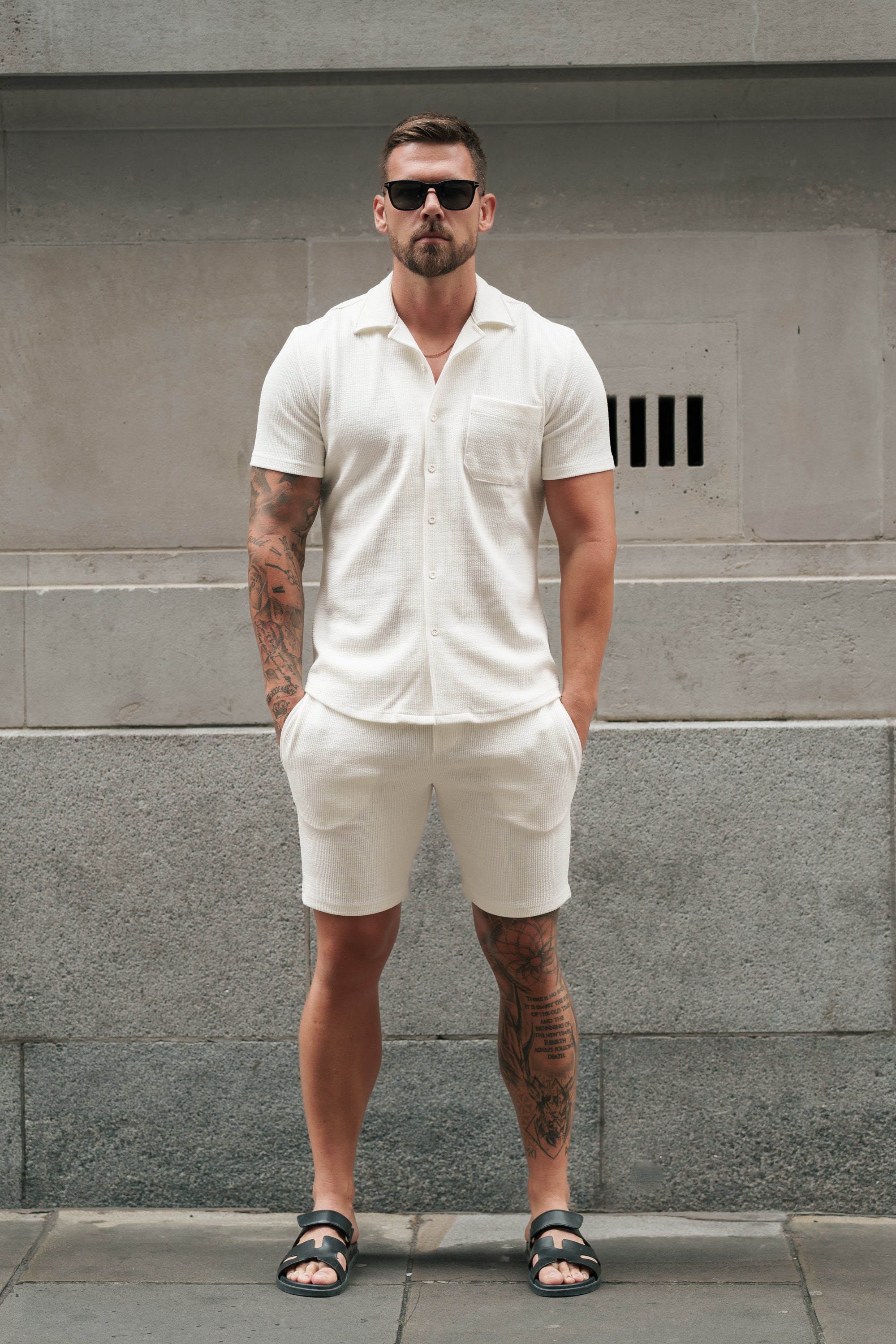 Man wearing a white shirt and shorts standing against a gray wall.
