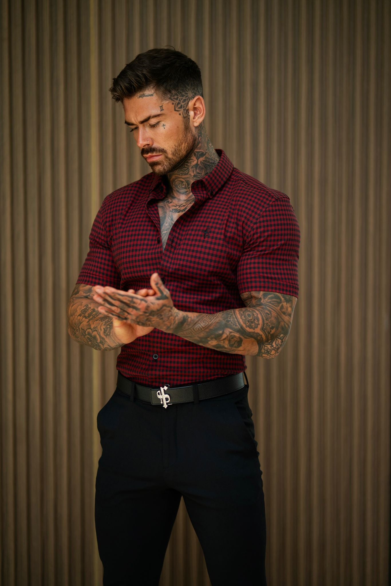 A man wearing a red and black check short sleeve shirt with a slim fit, standing in front of a striped background.