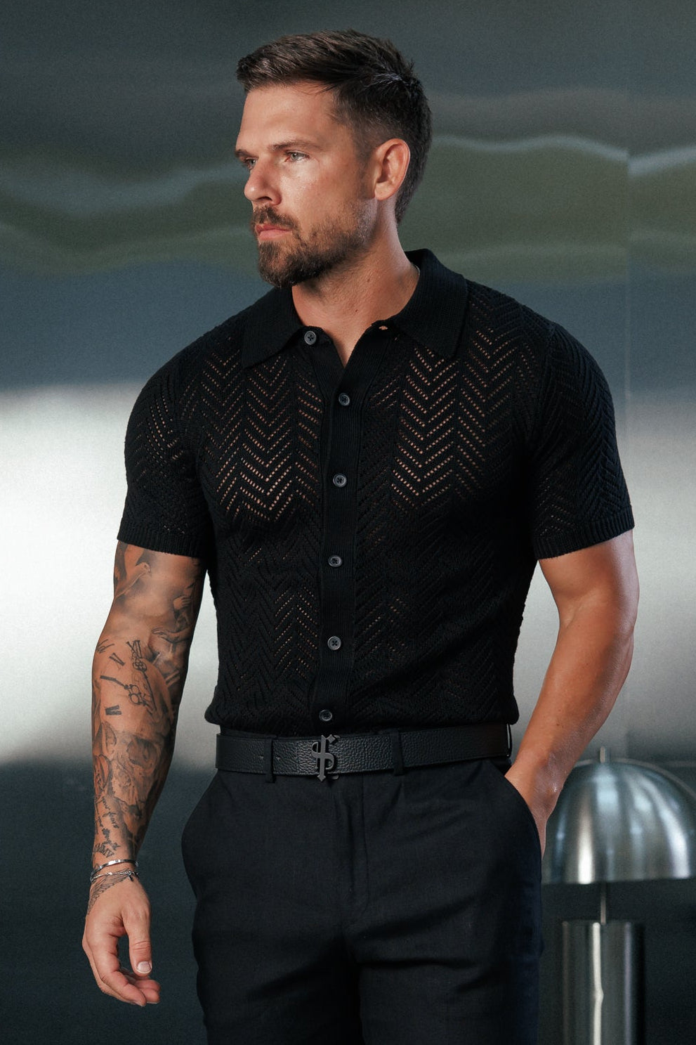 A man modeling a black short-sleeve button-up shirt with textured knit details on the chest and sleeves, paired with black trousers and black leather loafers, standing in a modern, dimly lit room with a metallic wall.