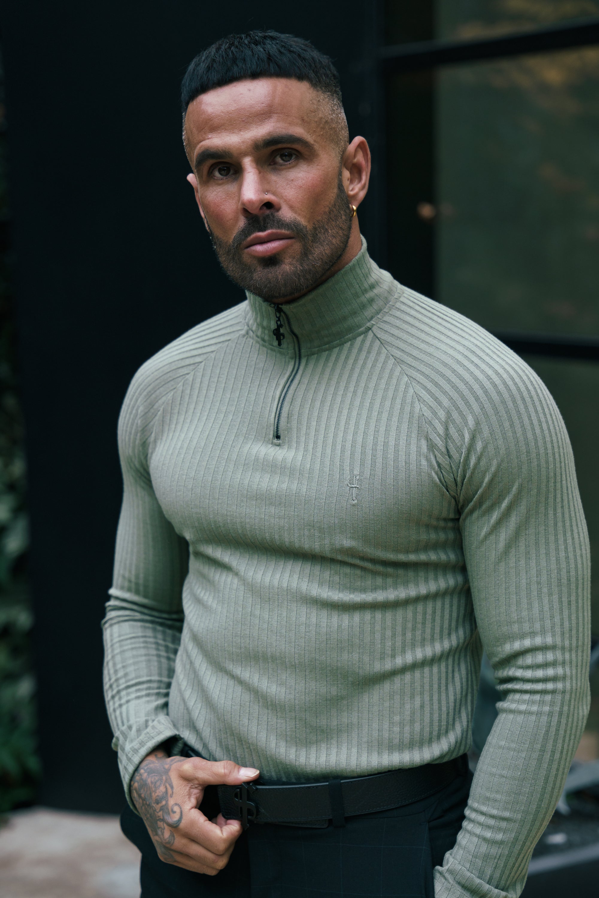 A male model wearing an olive green ribbed zip funnel neck raglan long sleeve crew neck jumper.