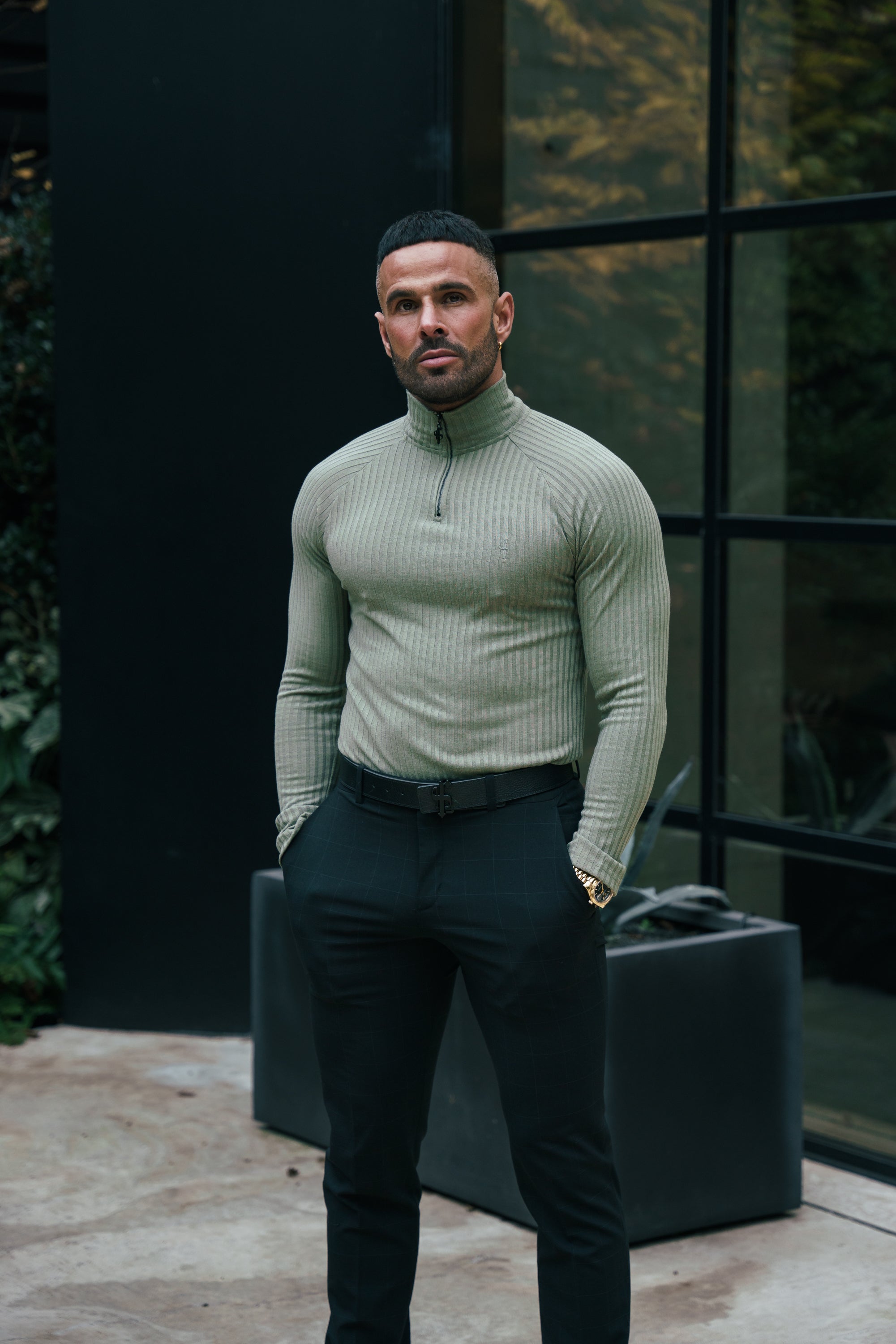 A man wearing an olive green ribbed zip funnel neck raglan long sleeve crew neck sweater and black trousers standing outdoors.