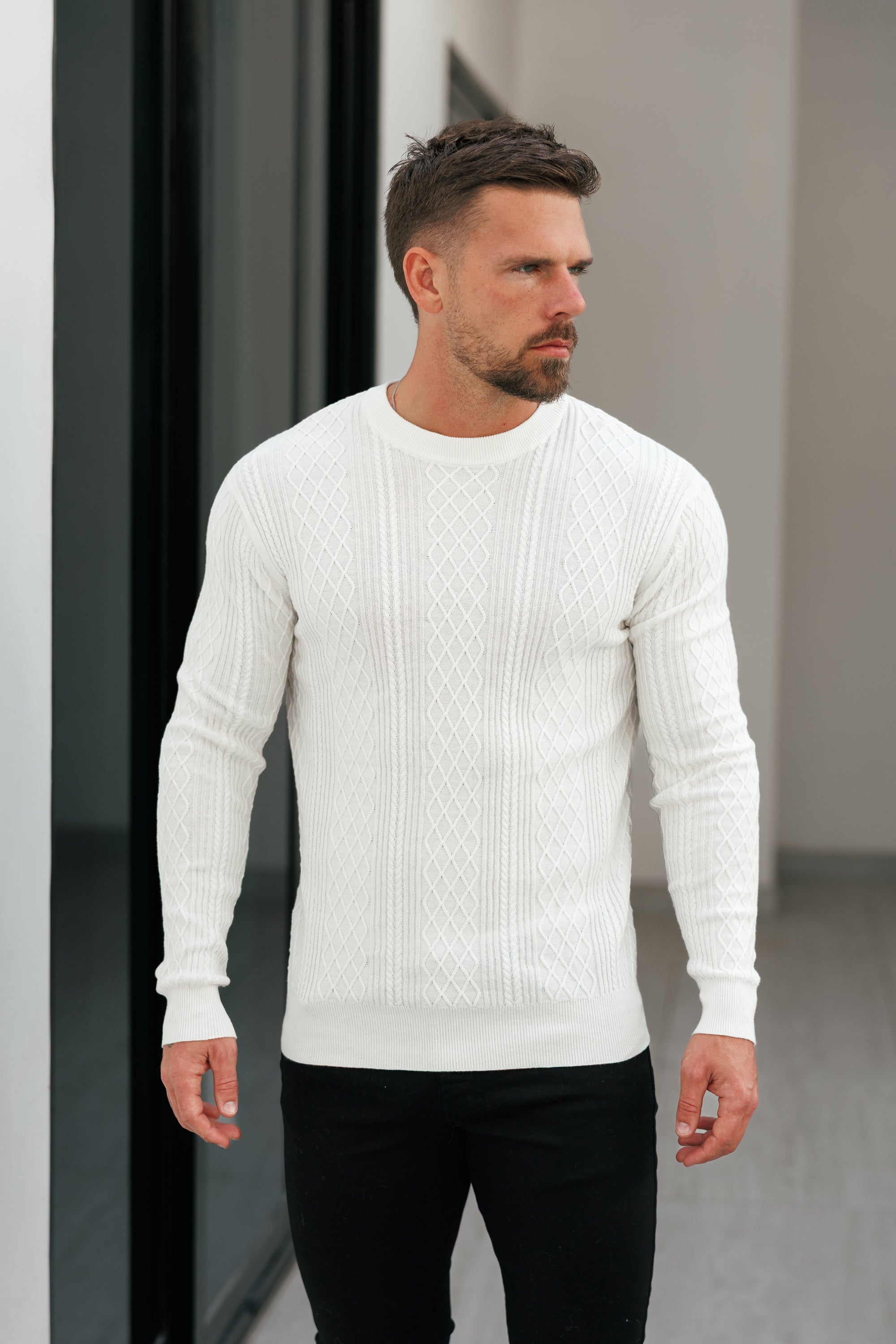 Man wearing a white sweater and black pants standing against a neutral background