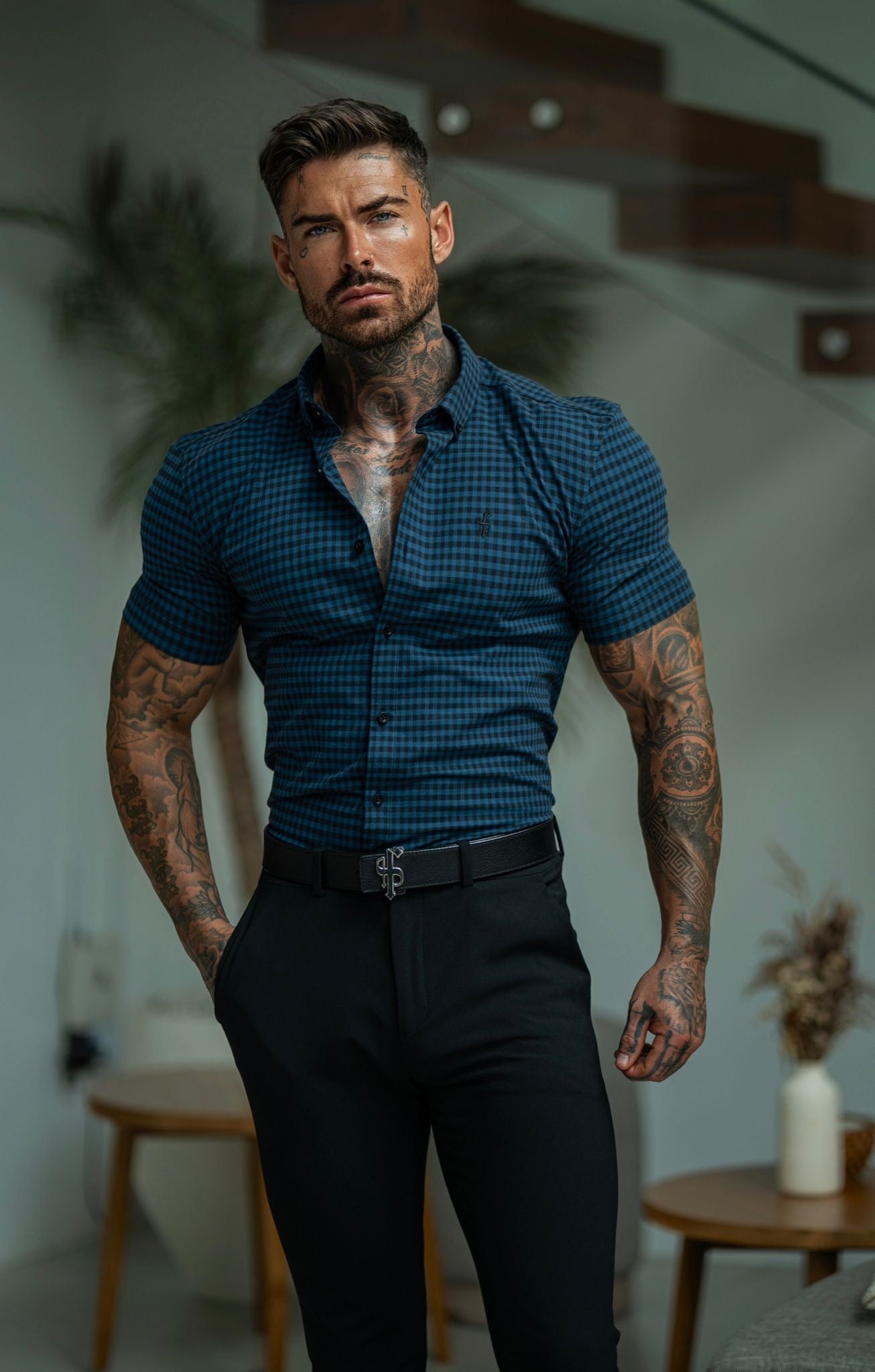 A man wearing a short sleeve blue and black check shirt with black trousers. The shirt features a super slim fit and the Father Sons tonal black emblem.
