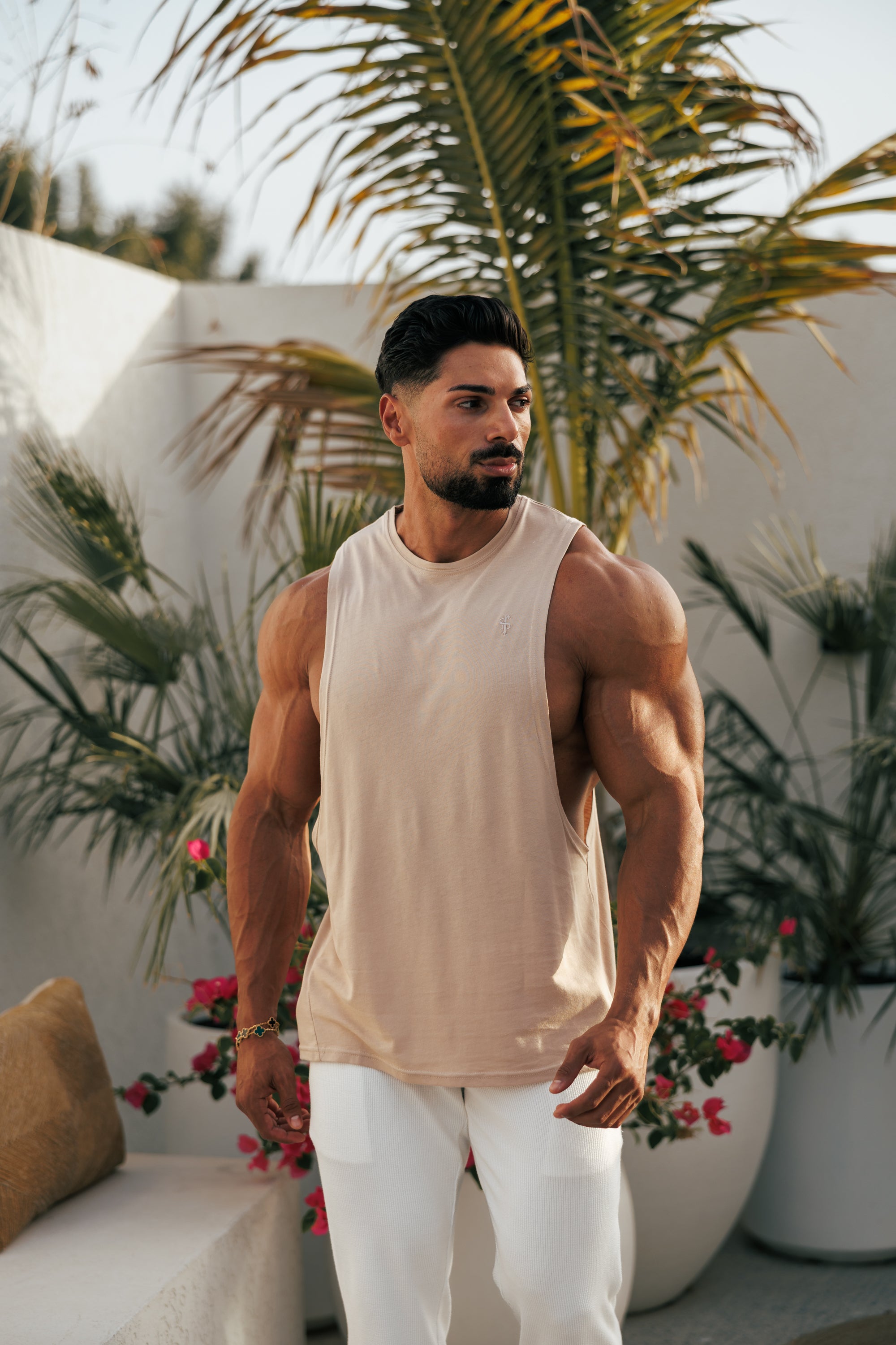 A male model wearing a beige sleeveless vest with a round neckline, paired with white trousers, standing outdoors with tropical plants in the background.