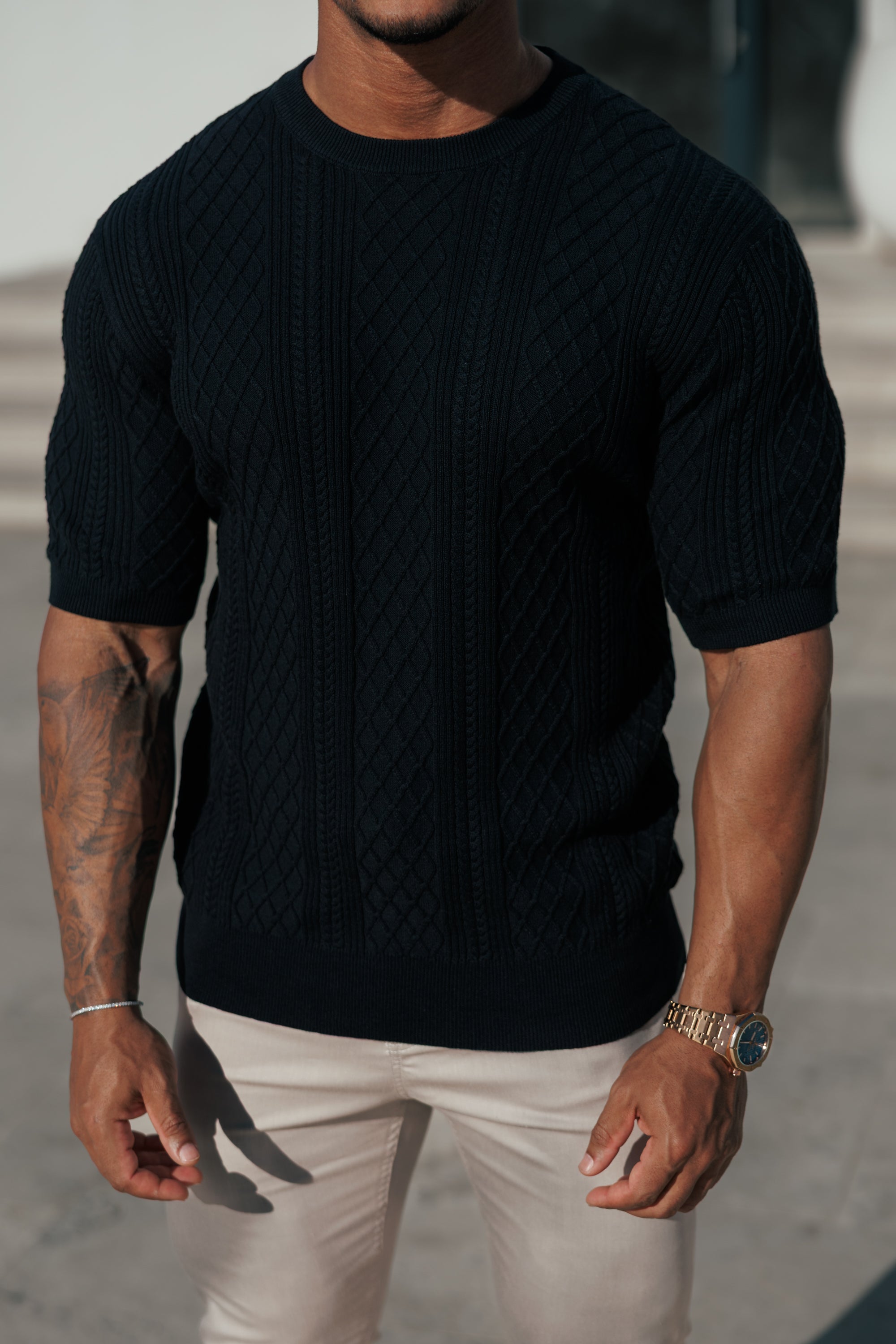 Person wearing a black ribbed sweater and white pants with tattoos on arms.